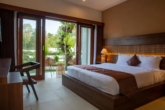 A bedroom with a king bed, a work desk, and sliding glass doors opening onto a terrace with rattan chairs and a garden.