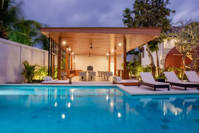A swimming pool at dusk with sun loungers along its edge and a covered open-sided pavilion with dining tables and ceiling fans behind it.