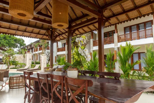 A covered open-air dining pavilion with a long wooden table and cross-back chairs, with a pool and sun loungers visible at left and guest rooms with balconies behind.