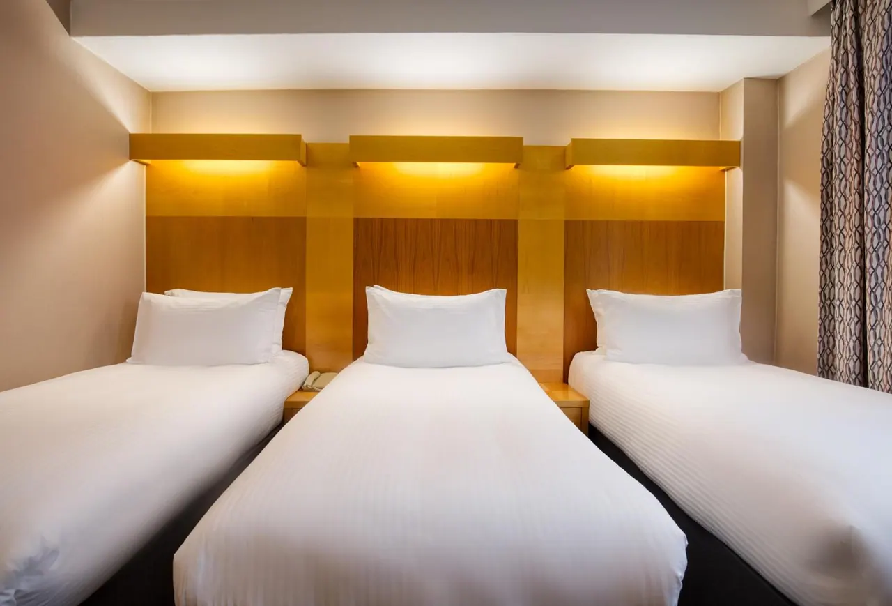 Three single beds with white bedding stand against a wooden headboard wall lit by warm wall lights, with a telephone on a bedside table.