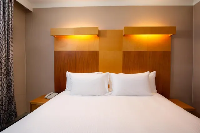 A made bed with white linen and four pillows sits against a wooden headboard with warm backlighting, with a bedside table and telephone visible on the left.
