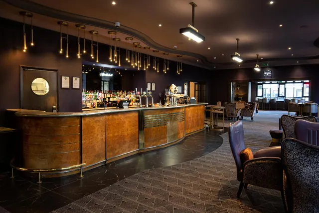 A lounge bar with a curved wooden counter, shelves of bottles, pendant lights and upholstered armchairs.