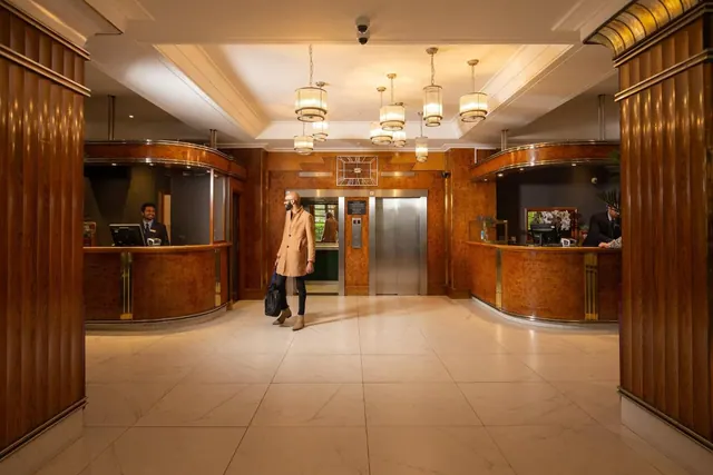 A lobby with wood-panelled columns, two reception desks with staff, pendant lights, a lift, and a person in a camel coat walking across a tiled floor.
