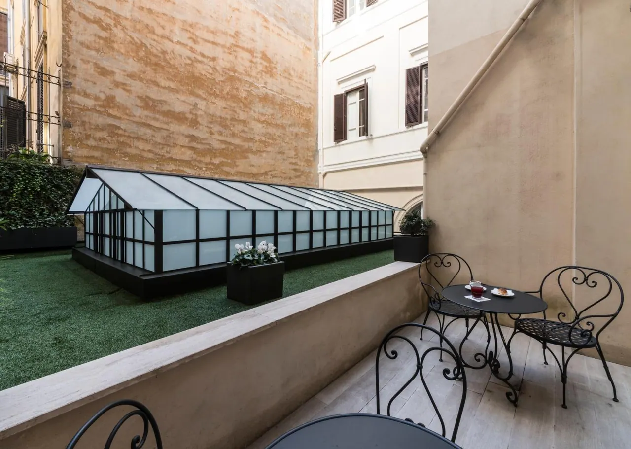 A furnished terrace with wrought iron chairs and a round table overlooks an enclosed courtyard with artificial grass, a glass-roofed structure and potted plants.