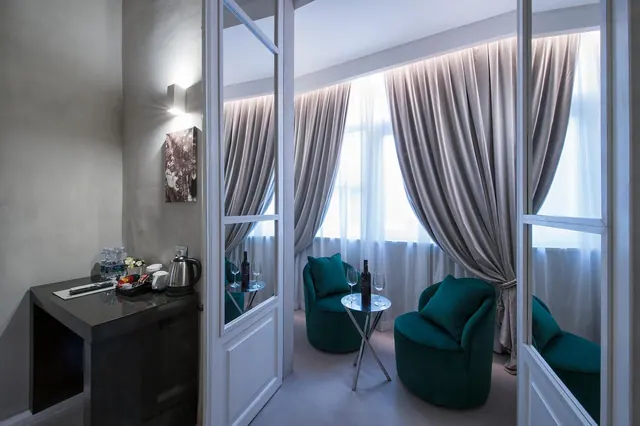 A sitting area with two teal armchairs and a round table holding a wine bottle and glasses, with grey curtains at a tall window, seen through glass doors, and a kettle and water bottles on a side unit to the left.