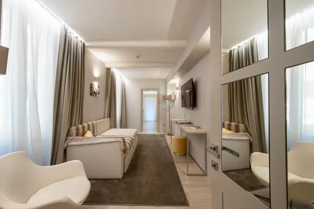 A long, narrow hotel room with a white sofa bed, a white armchair in the foreground, a wall-mounted television, a desk with a stool, and a mirrored wardrobe door on the right.