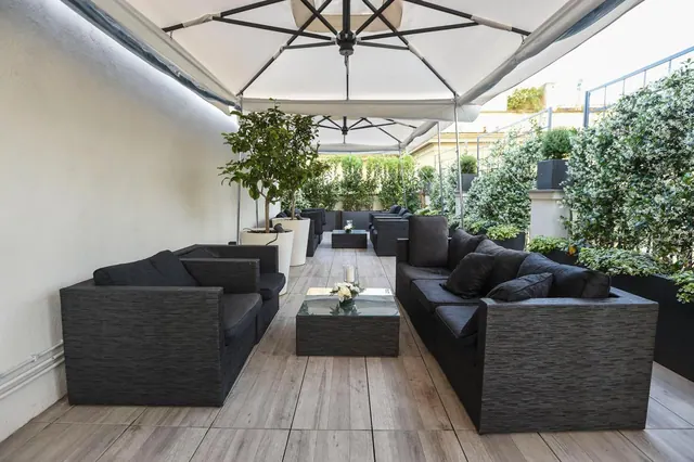 A covered outdoor terrace with dark wicker sofas, a glass coffee table with white flowers, potted trees and large white canopy umbrellas.