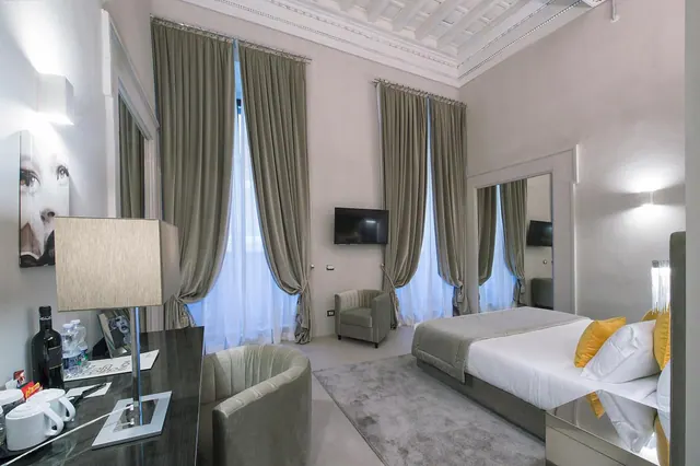 A bedroom with a double bed, tall curtained windows, a wall-mounted television, an armchair and a writing desk with a lamp and drinks.
