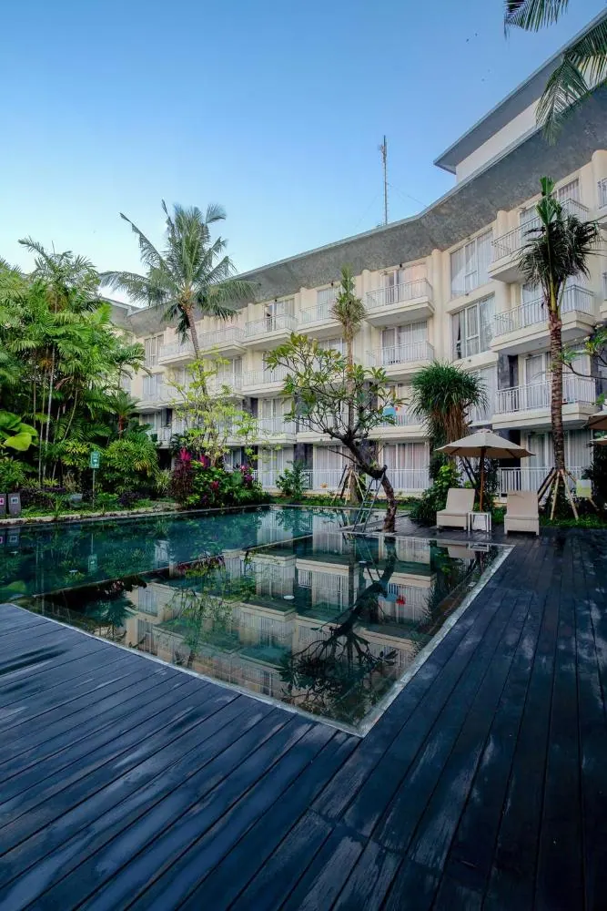 A rectangular swimming pool with a dark timber deck, two sun loungers and a parasol, in front of a multi-storey hotel building with balconies and palm trees.