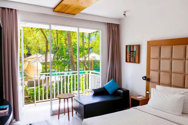 A bedroom with a double bed, a black leather chaise with a blue cushion, and a sliding glass door opening onto a balcony overlooking trees and a pool with umbrellas.