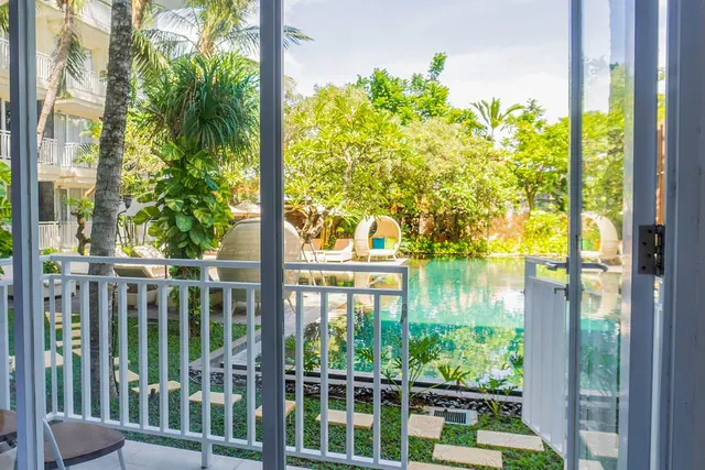 View through glass doors from a balcony with a white railing over a turquoise pool, sun loungers and wicker egg chairs in a tropical garden.