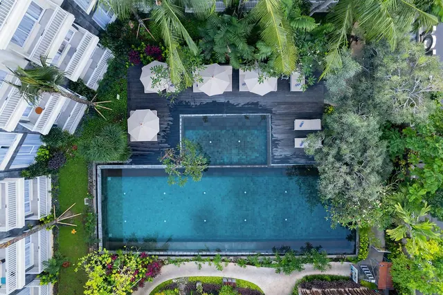 Aerial view of a swimming pool with a long rectangular section and a smaller pool on a timber deck, with white umbrellas, sun loungers, palm trees and villas around it.