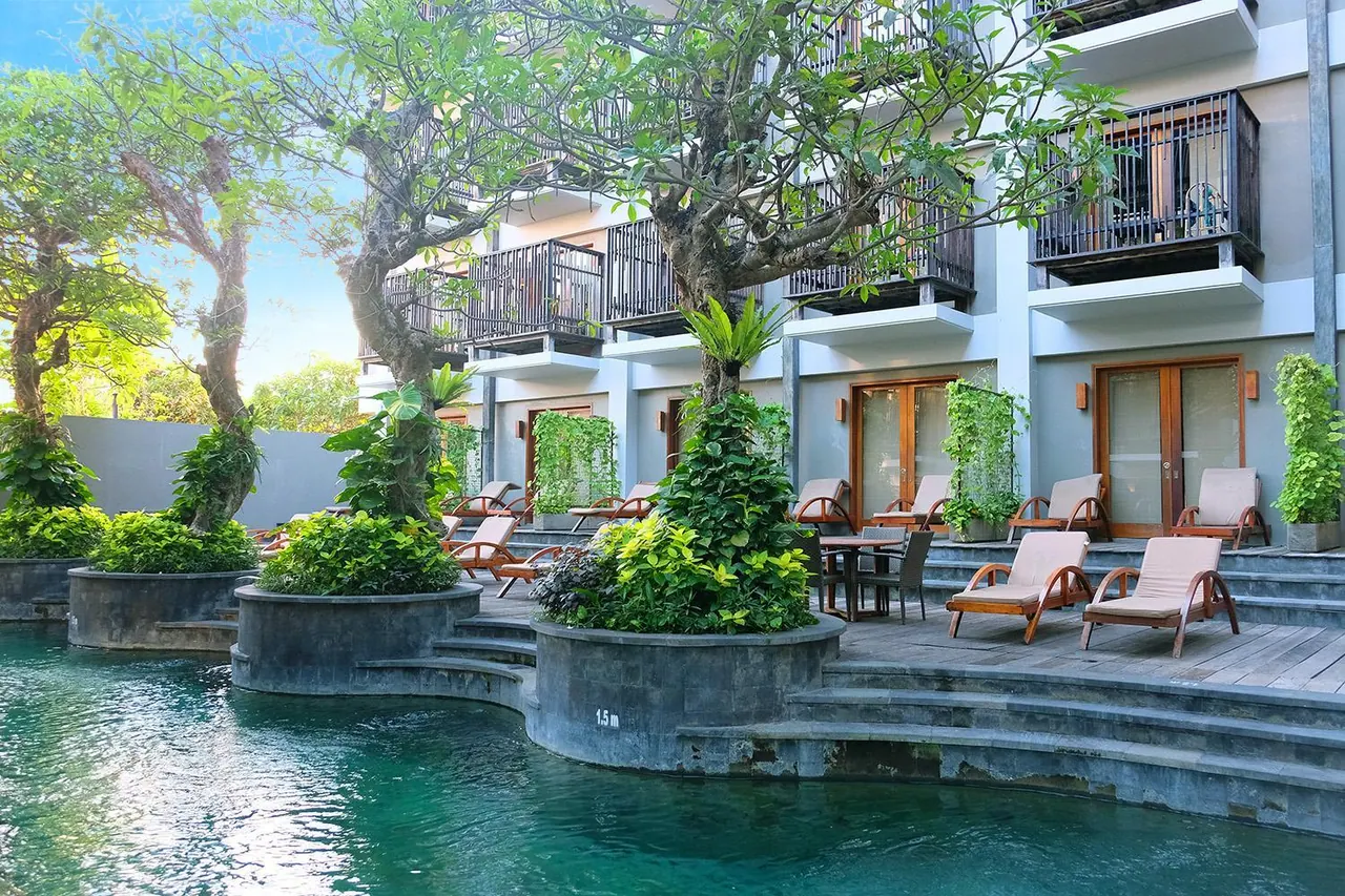 A pool with a stepped stone edge, timber loungers on a deck, and a multi-storey building with balconies behind trees.