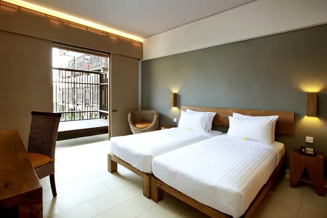A hotel bedroom with two beds made with white linens, a wicker armchair, a wooden desk and chair, and a sliding door opening onto a balcony that faces a neighbouring building.
