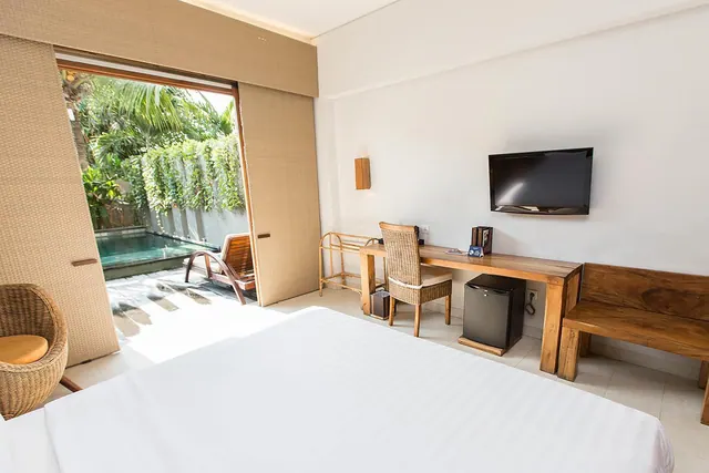 A bedroom with a white bed, a wooden desk and wicker chair, a wall-mounted television, and sliding woven doors opening onto a terrace with a small pool, a wooden lounger and tropical plants.