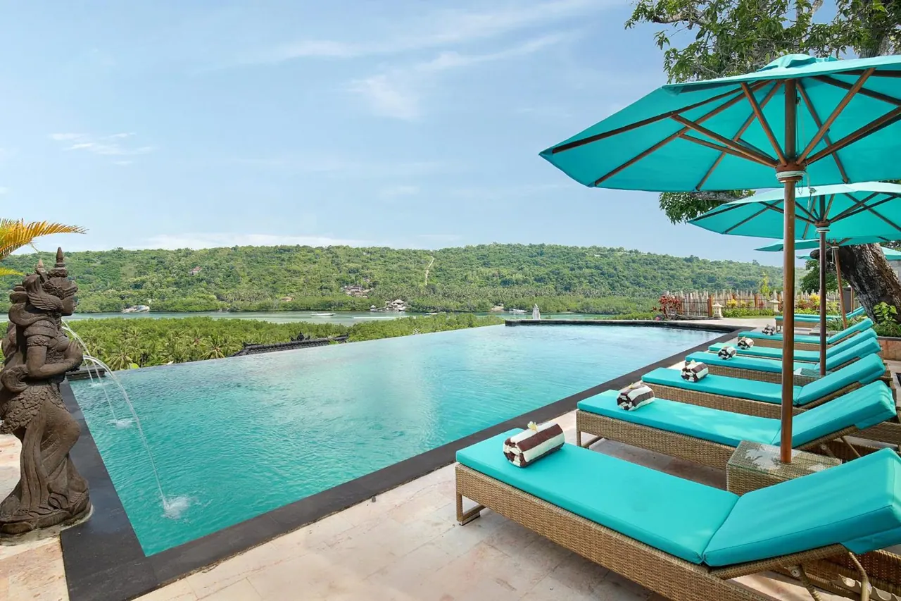 An infinity pool with teal-cushioned sun loungers and teal umbrellas overlooks a bay and wooded hills, with a stone statue spouting water at the left edge.