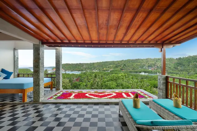A covered terrace with a wooden ceiling, checkerboard floor, two loungers with teal cushions, and a petal heart design on the floor, looking out over green hills and a bay.