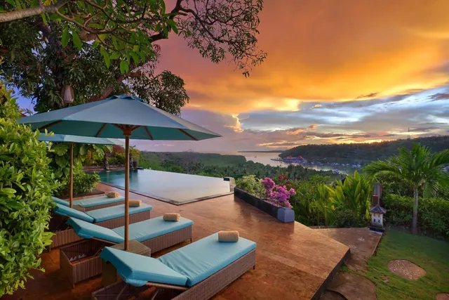 An infinity-edge pool, sun loungers and parasols on a terrace overlooking a bay at sunset.