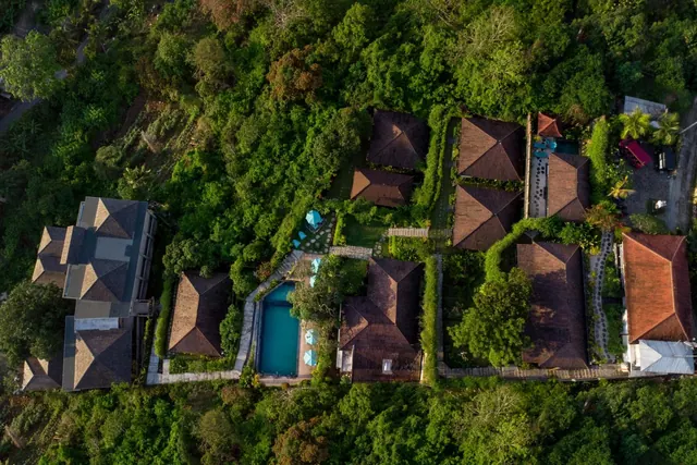 Aerial view of a resort with tiled-roof villas, a small rectangular pool with blue umbrellas, and paths set among dense jungle trees.