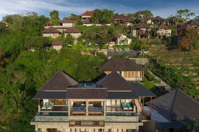 Aerial view of a hillside resort with thatched-roof villas, a pool with umbrellas, and balconies with loungers overlooking dense green jungle.