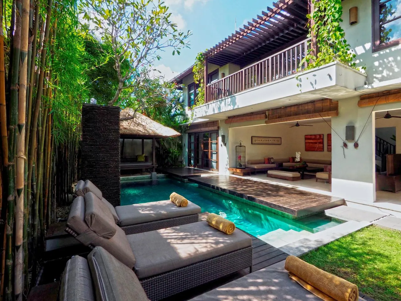 A private swimming pool with sun loungers and rolled towels sits beside a two-storey villa with a covered lounge area and an upstairs balcony.