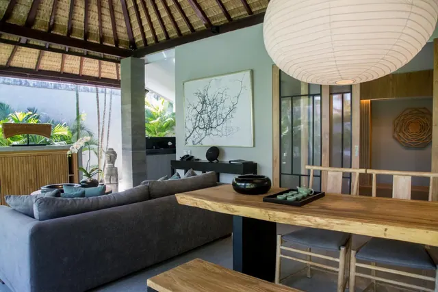 A living area with a grey sofa, a live-edge wooden dining table and a large paper lantern, with a garden visible through the open thatched pavilion.
