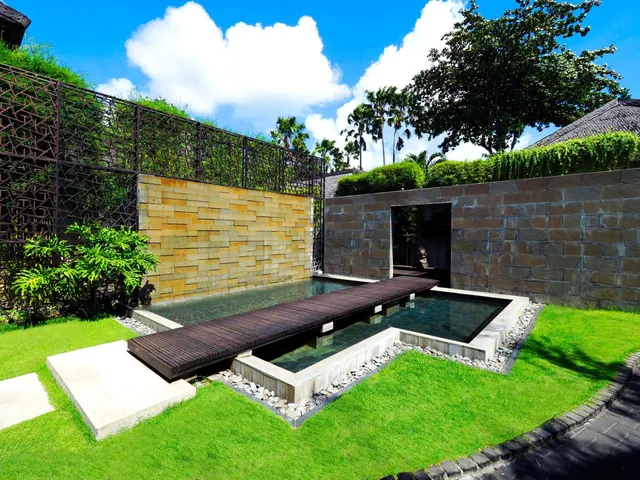 A small rectangular pool with a timber walkway across it sits in a lawned garden beside a stone wall and a latticed screen.