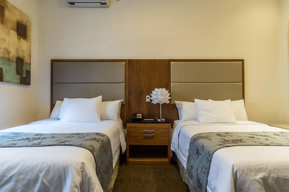 Two twin beds with white bedding and upholstered grey headboards sit either side of a wooden nightstand holding a lamp and telephone, with a wall-mounted air conditioner above.