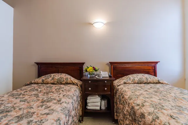 Two single beds with wooden headboards and floral bedspreads sit either side of a nightstand holding a flower arrangement and a telephone.
