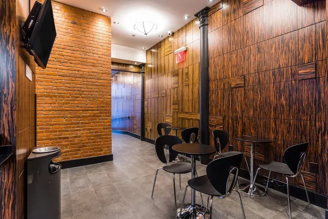 A seating area with small round tables and black chairs sits beside an exposed brick wall and a wood-panelled wall, with a mounted television and an exit sign visible.