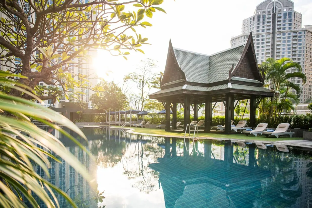 An outdoor pool with sun loungers, shade umbrellas and a traditional Thai-style pavilion beside the water, with tall buildings behind.
