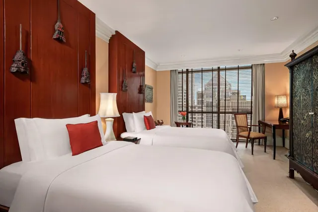 A hotel bedroom with two white beds, red cushions, a carved wooden headboard, a desk and armchair, and a window looking onto high-rise buildings.