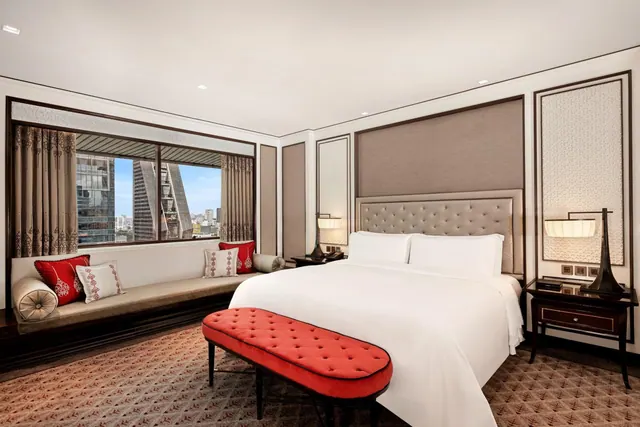 A hotel bedroom with a king bed, a red tufted bench at the foot, and a window seat overlooking high-rise buildings.