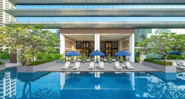 A large swimming pool in front of a hotel building, with blue umbrellas, sun loungers and frangipani trees in planters along the deck.