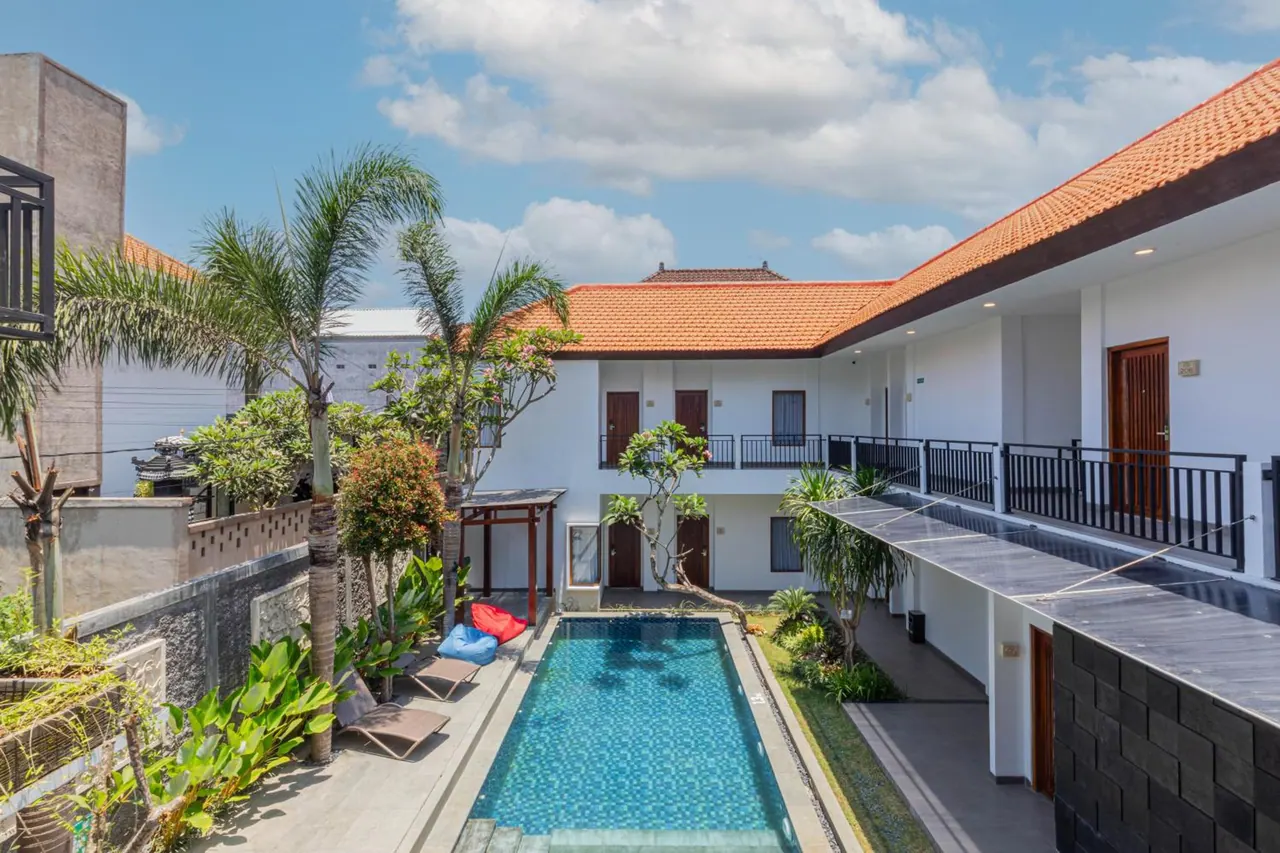 A long rectangular swimming pool sits in a courtyard lined with sun loungers, palm trees and two-storey white buildings with orange tiled roofs.