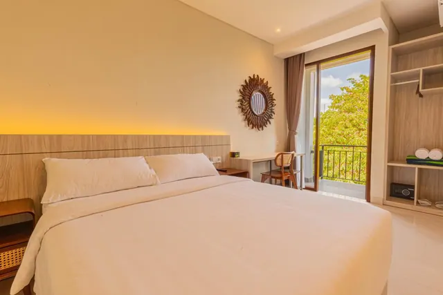 A bedroom with a made double bed, a backlit wooden headboard, a desk and chair, an open wardrobe, and a balcony door looking onto trees.
