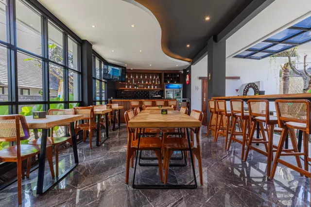 A restaurant area with wooden tables and cane chairs on a dark marble floor, a bar with bottles and high stools, and tall windows showing greenery outside.