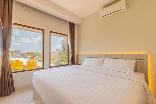 A bedroom with a made king bed, a wall-mounted air conditioner, and two windows looking out over rooftops and trees.