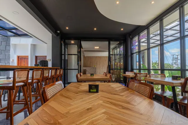 A restaurant dining room with a herringbone wooden table, rattan chairs and bar stools, and floor-to-ceiling windows showing banana plants outside.