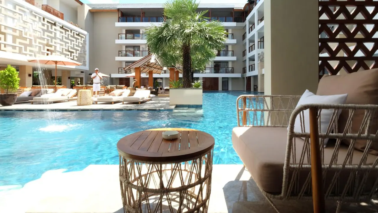 A large swimming pool with sun loungers, a shaded umbrella and a palm tree, with a rope lounge chair and round side table in the foreground.
