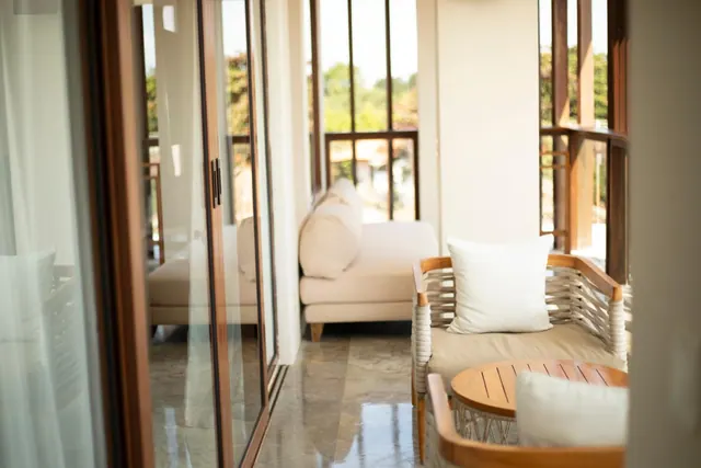 A covered terrace with wicker armchairs, cushions and a round wooden table, with a sofa further back and trees visible through the glass doors.