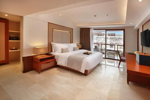A staged bedroom with a king bed, a wooden desk with a wall-mounted television, and sliding glass doors to a balcony overlooking a neighbouring patterned building.