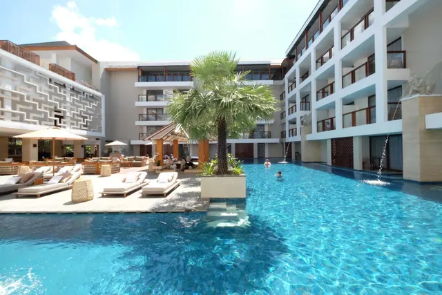A large outdoor swimming pool with a palm planter in the middle, surrounded by sun loungers, umbrellas and hotel buildings, with two swimmers in the water.