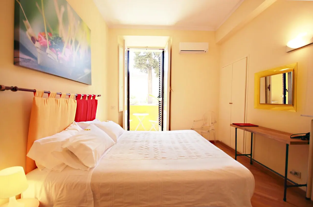 A bedroom with a double bed, yellow walls, a wall-mounted air conditioner, a desk and mirror, and an open door to a small balcony with a table and chairs.