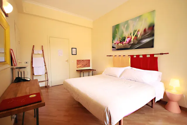 A bedroom with a double bed with white linen, a red and yellow headboard drape, a wooden desk, a wooden towel ladder and a pink side table with a lamp.