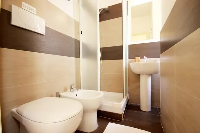 A bathroom with a wall-hung toilet, a bidet, a pedestal sink and a glass-enclosed shower, with brown and beige tiled walls.