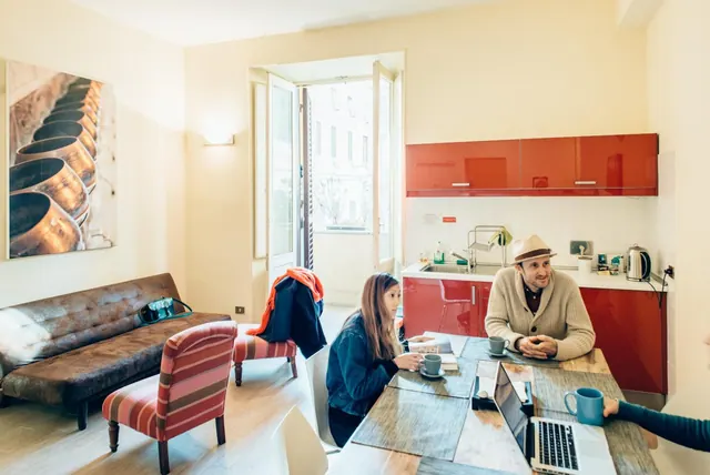 A combined living and dining area with a red kitchenette along the back wall, a brown leather sofa and striped armchairs, a dining table where two people sit, and open French doors onto a street.