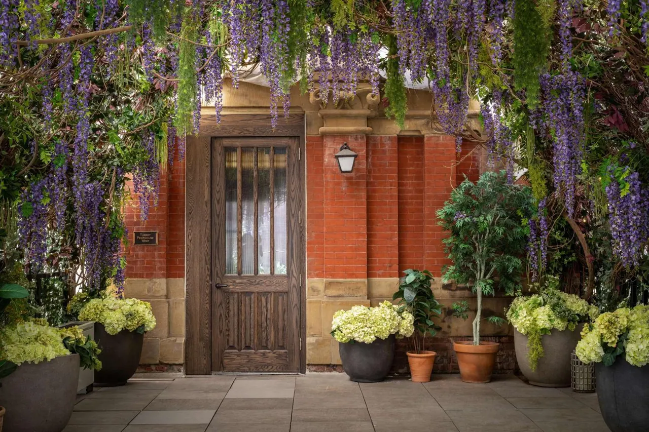 A wooden door with glass panes is set in a red brick wall, framed by overhead wisteria and potted hydrangeas and trees on a paved terrace.