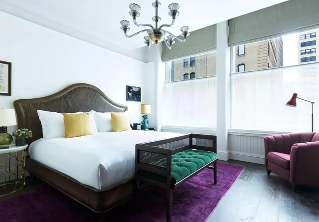 A bedroom with a king bed, a green tufted bench on a purple rug, a pink armchair, and tall windows looking out onto city buildings.