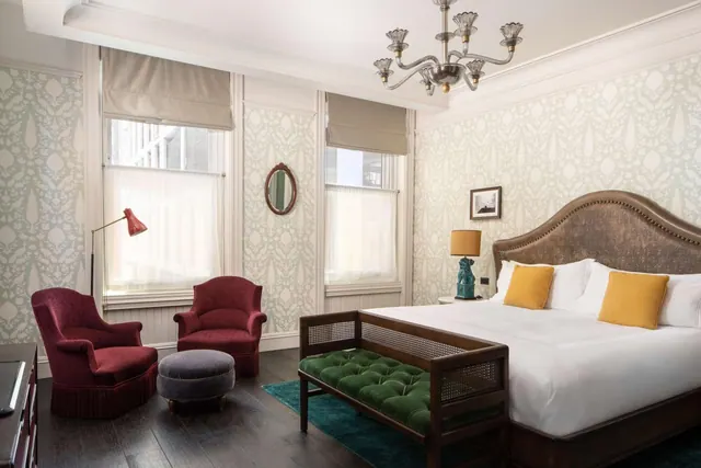 A bedroom with a king bed under a chandelier, two red armchairs and a footstool by the windows, and a green velvet bench at the foot of the bed.